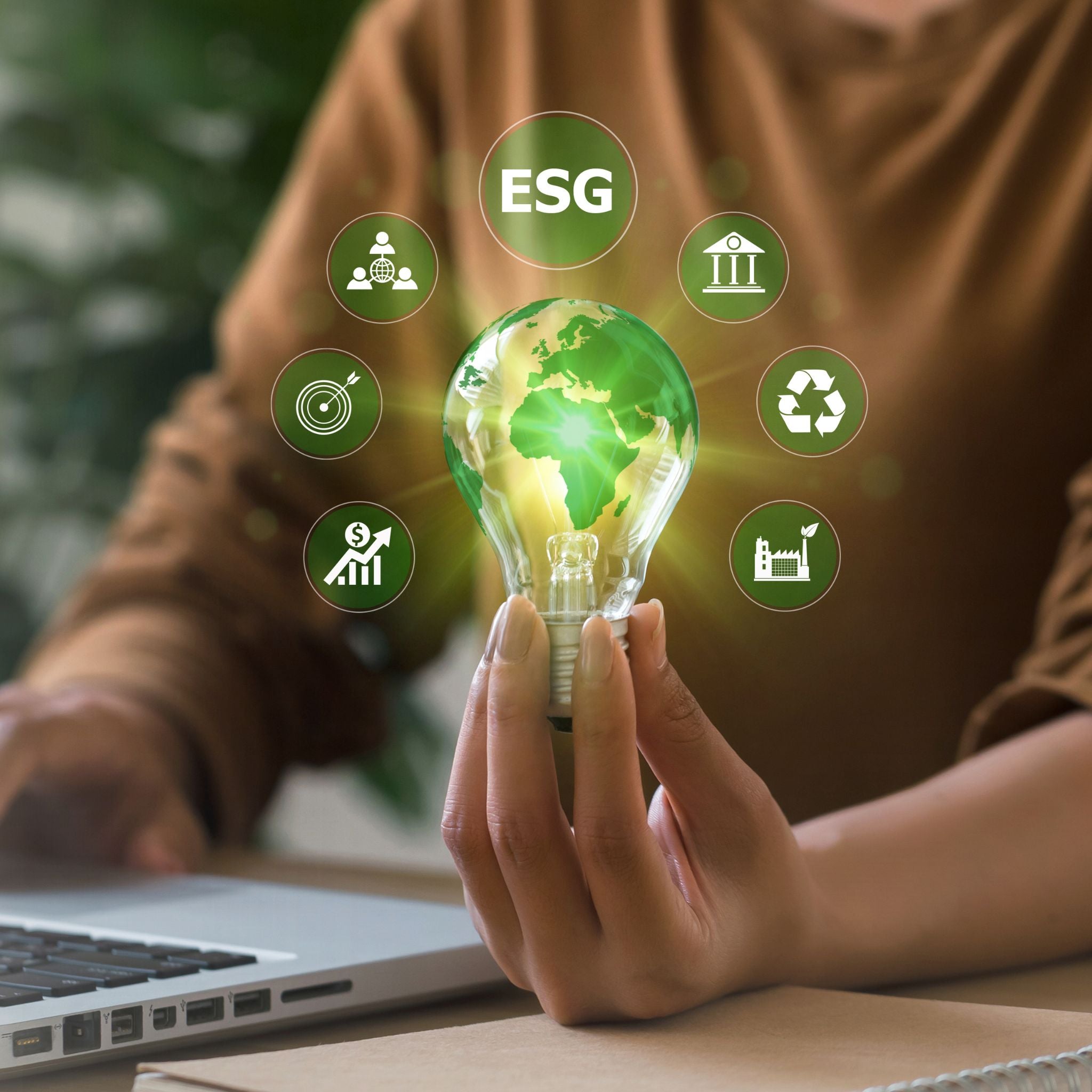 Person holding a light bulb with Earth design, surrounded by ESG icons, in front of a laptop.