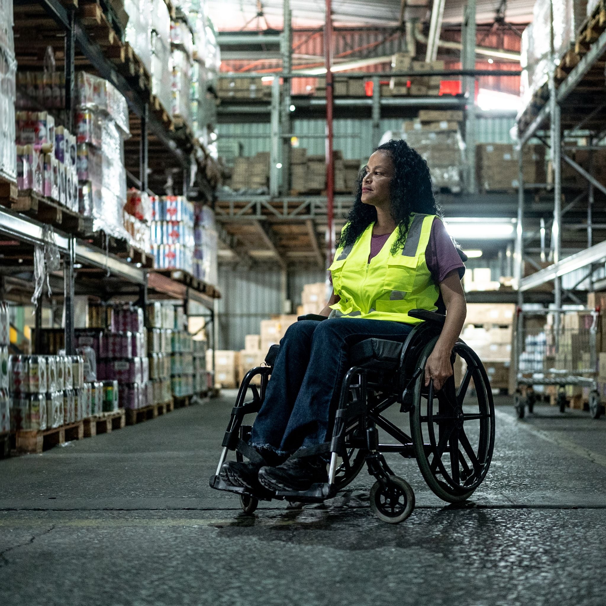 Person in a wheelchair wearing a high-visibility vest in a warehouse.