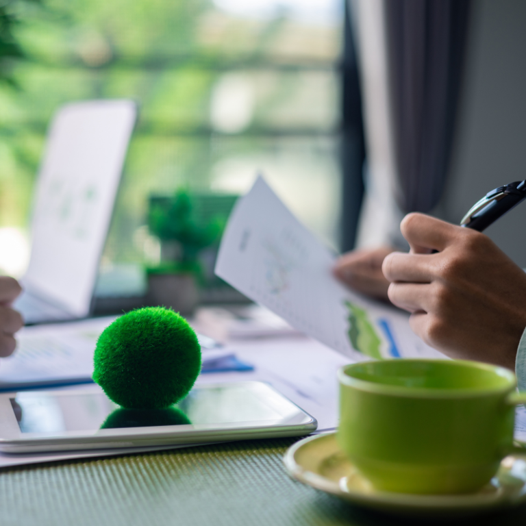 Two people working at a desk with a laptop, tablet, and green mug.